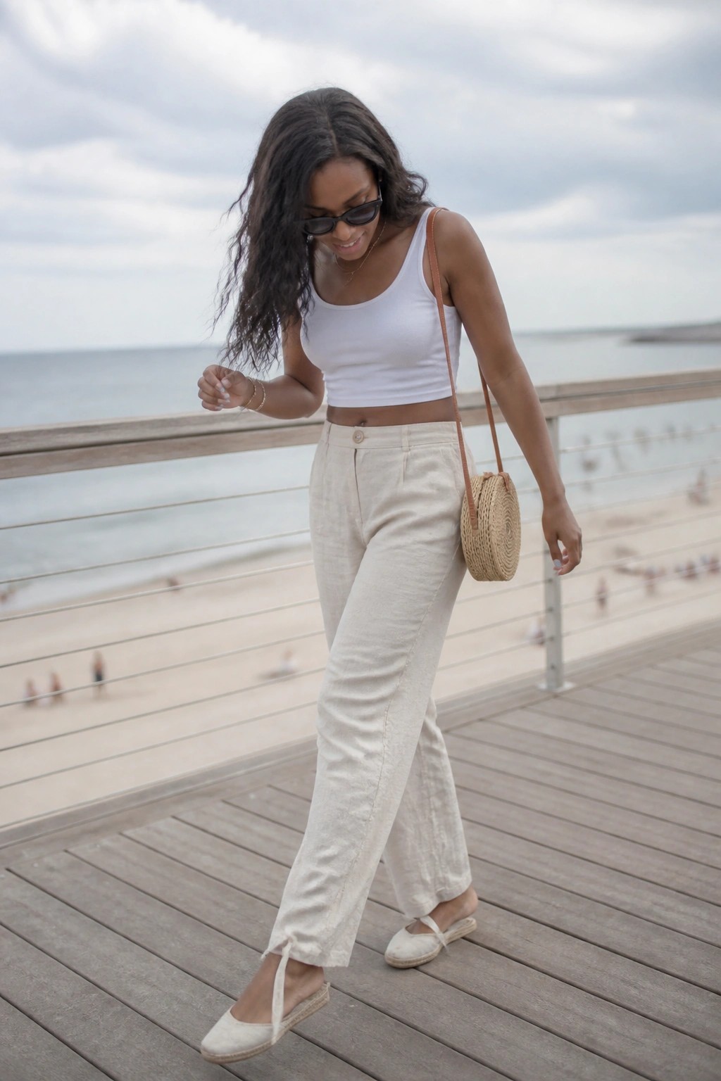 Woman in white crop top and beige linen pants on beach boardwalk with woven bag