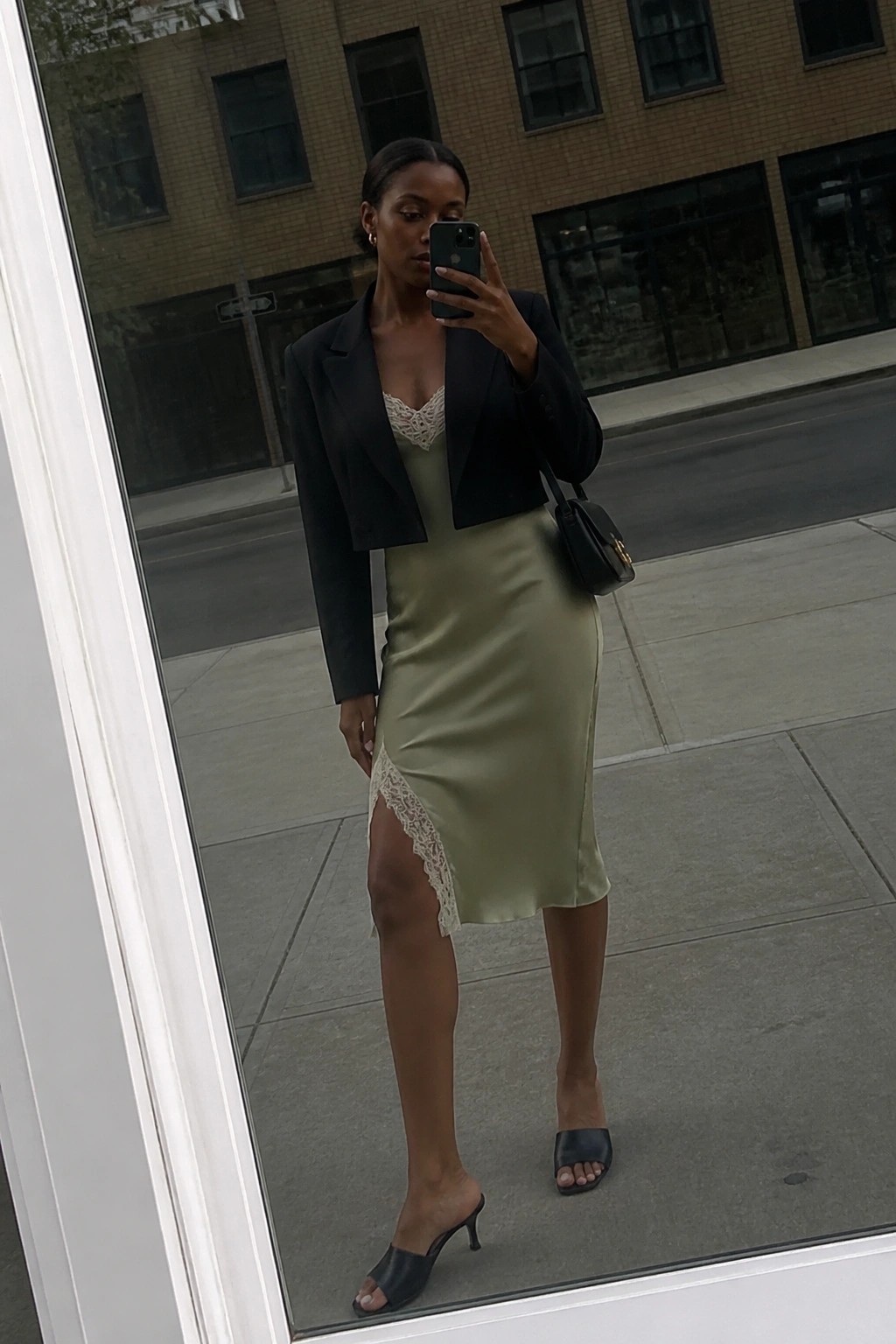 Woman wearing a cropped black blazer over a sage green satin slip dress with lace side slit detail, paired with black heeled mules and a small black handbag while taking a mirror selfie.