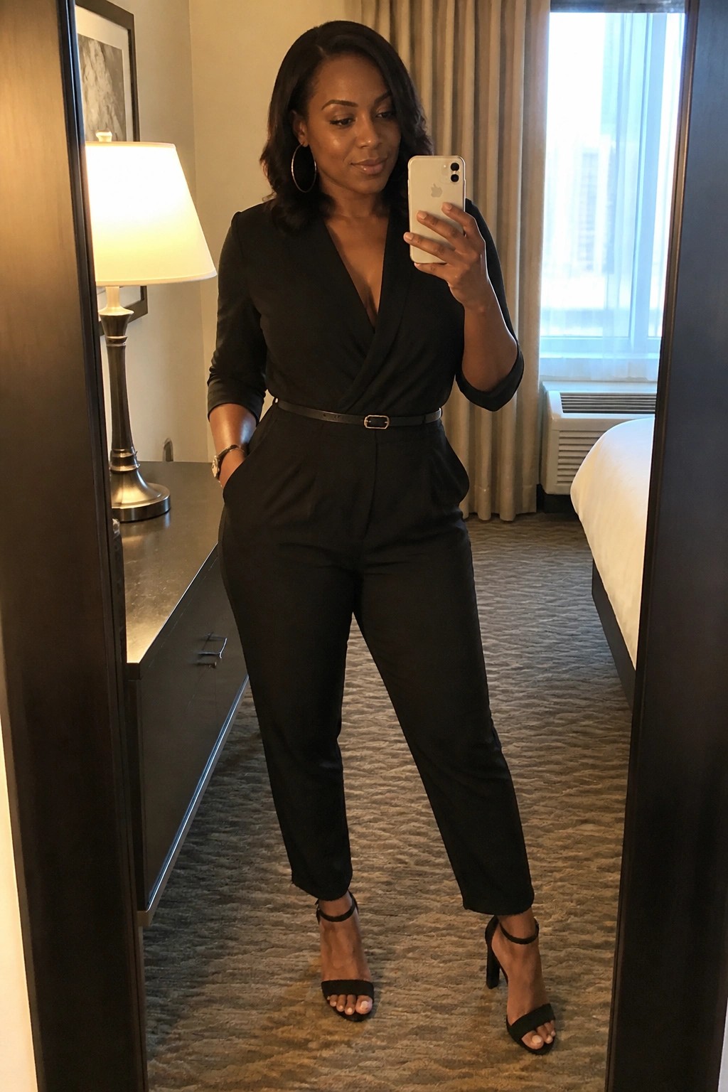 A woman in a black wrap jumpsuit with a thin belt three quarter sleeves and black strappy heels taking a mirror selfie indoors.