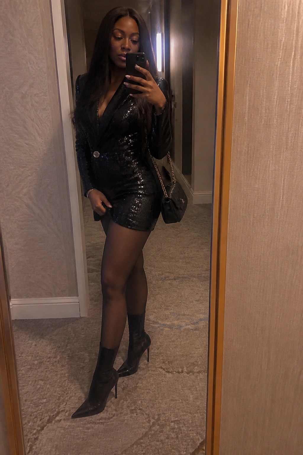 Woman wearing black sequined blazer mini dress with sheer tights, pointed stiletto boots, and black quilted chain strap bag in mirror selfie pose.