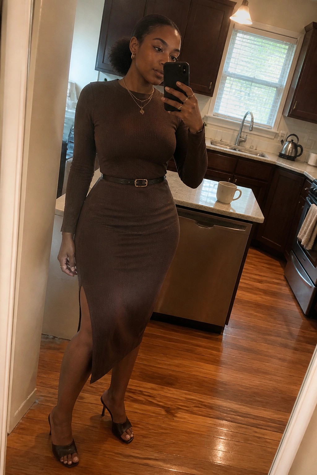 Woman wearing a brown ribbed midi dress with black belt gold necklaces and black heeled mules posing for a mirror selfie indoors