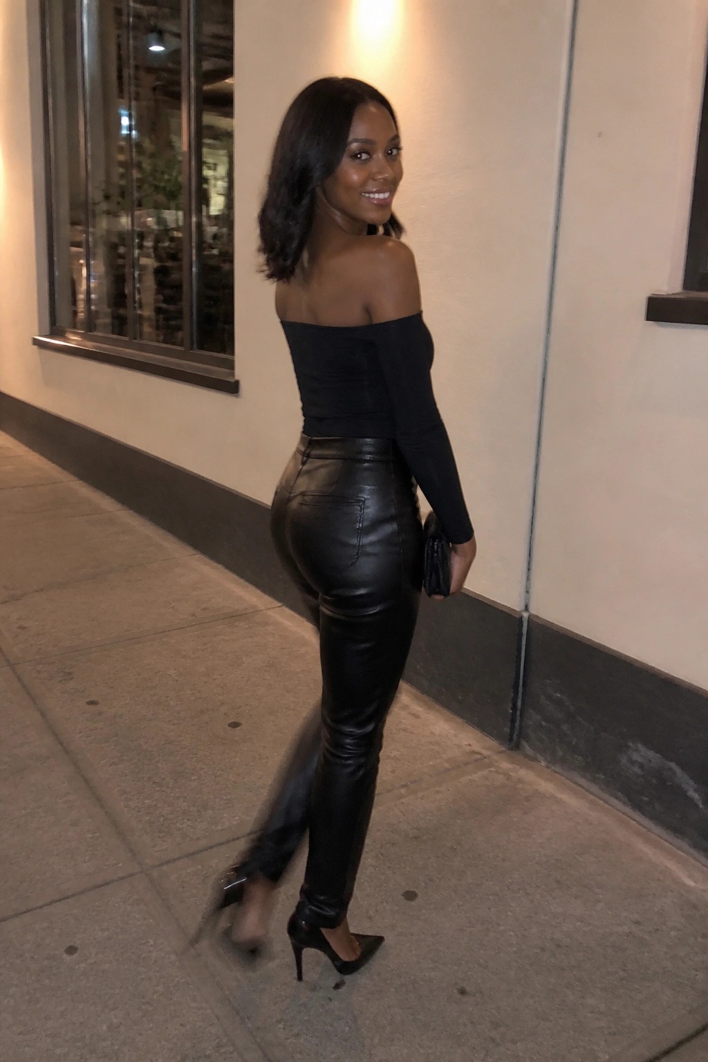 Woman wearing a fitted black off shoulder long sleeve top with black leather pants and pointed toe heels holding a small clutch while posing on a sidewalk.