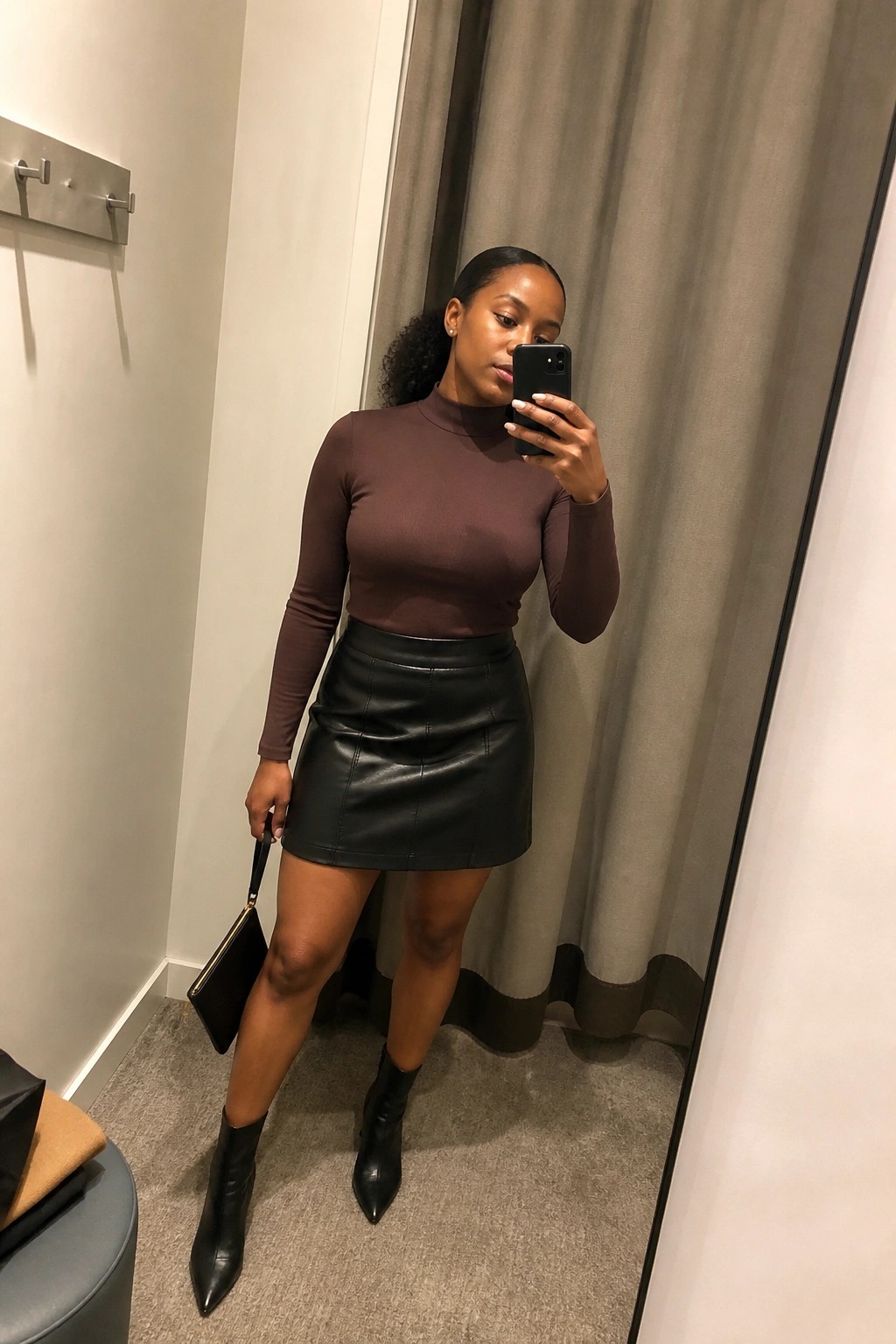 Woman in brown fitted turtleneck top and black leather mini skirt with black pointed ankle boots holding a black clutch in a mirror selfie.