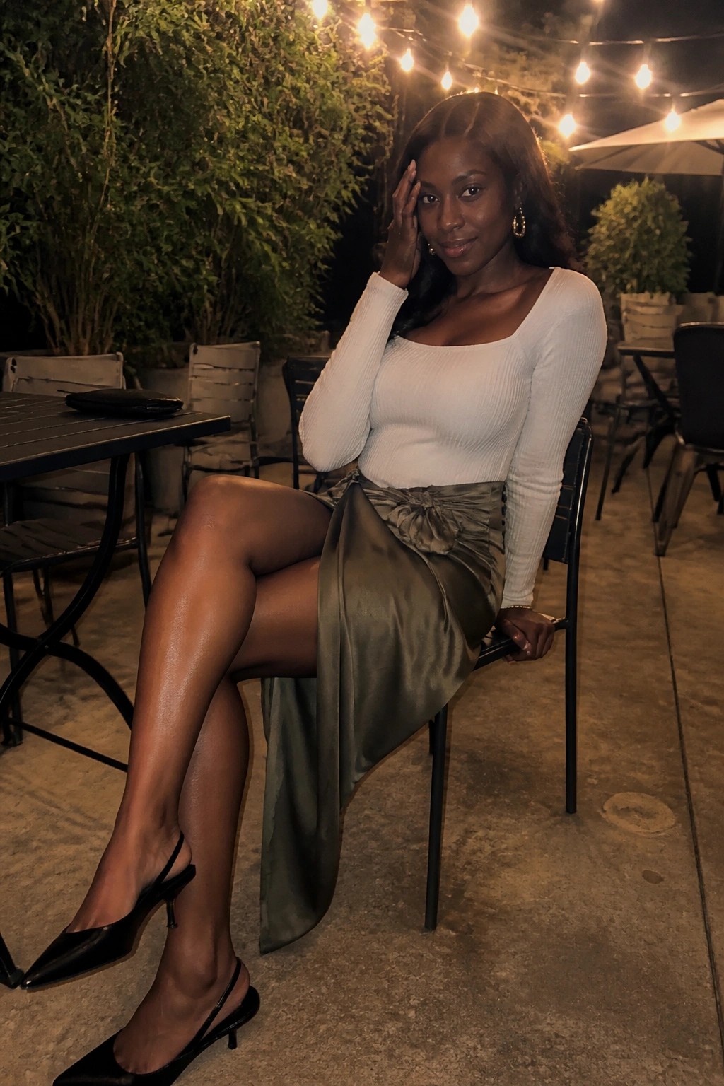 Woman in white ribbed top and olive satin skirt seated outdoors under string lights.