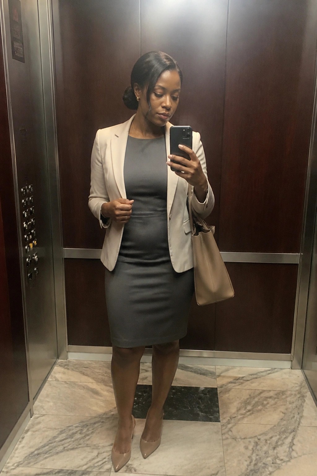 Woman in beige blazer and gray dress taking phone selfie in elevator
