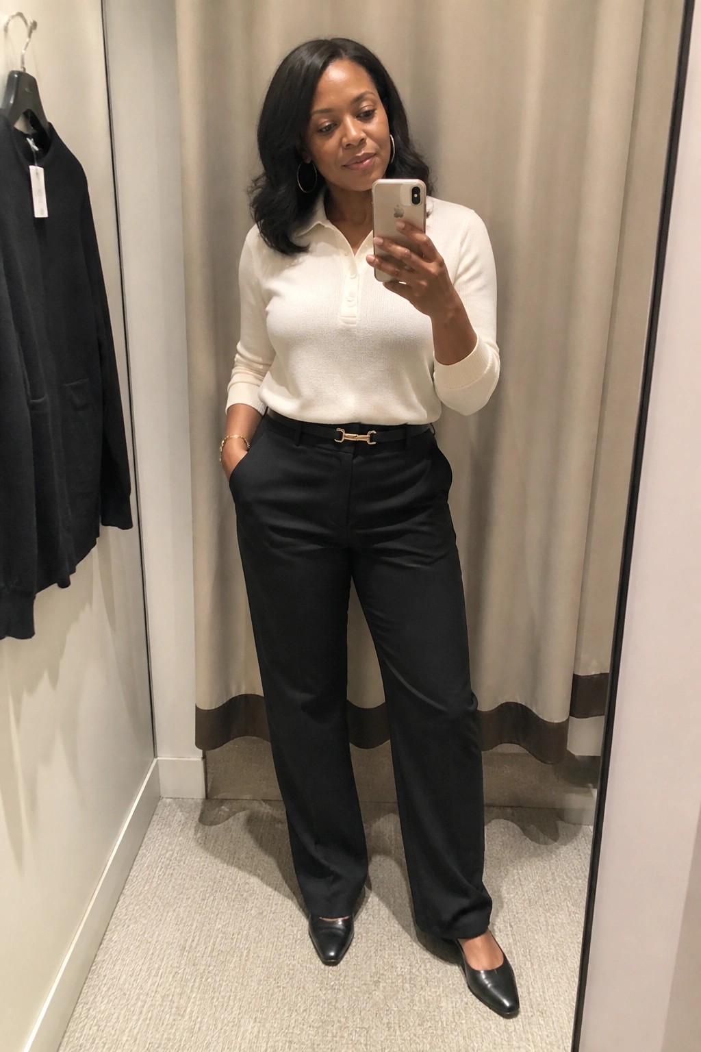 Woman taking a mirror selfie in a fitting room wearing a cream polo knit sweater tucked into black wide-leg trousers with a black belt featuring a gold buckle, paired with black pointed-toe flats and gold hoop earrings.