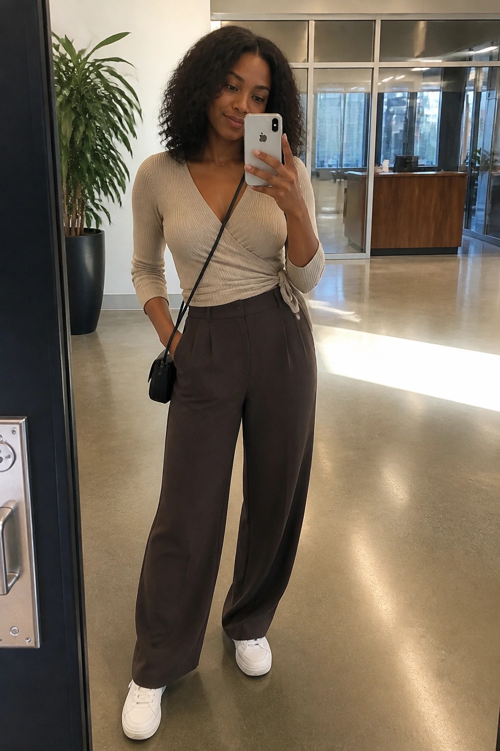 Woman in beige ribbed wrap top tied at side with brown wide leg trousers and white sneakers holding a phone in an office interior.