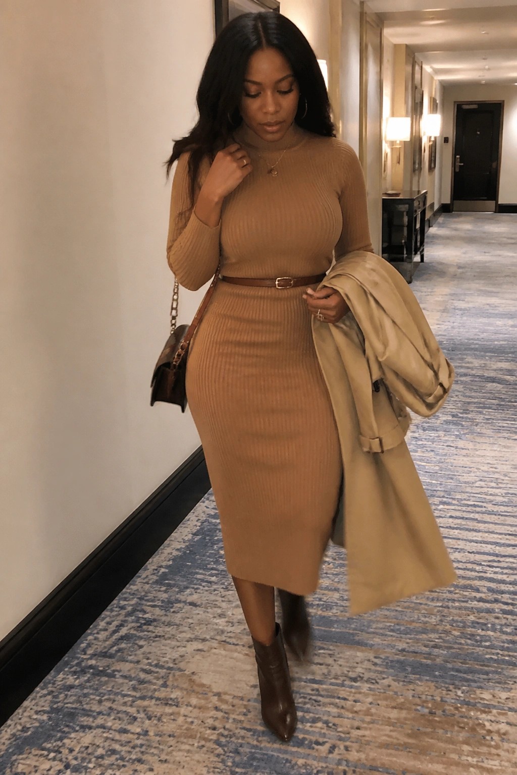 Woman in tan ribbed turtleneck dress holding beige trench coat in hallway