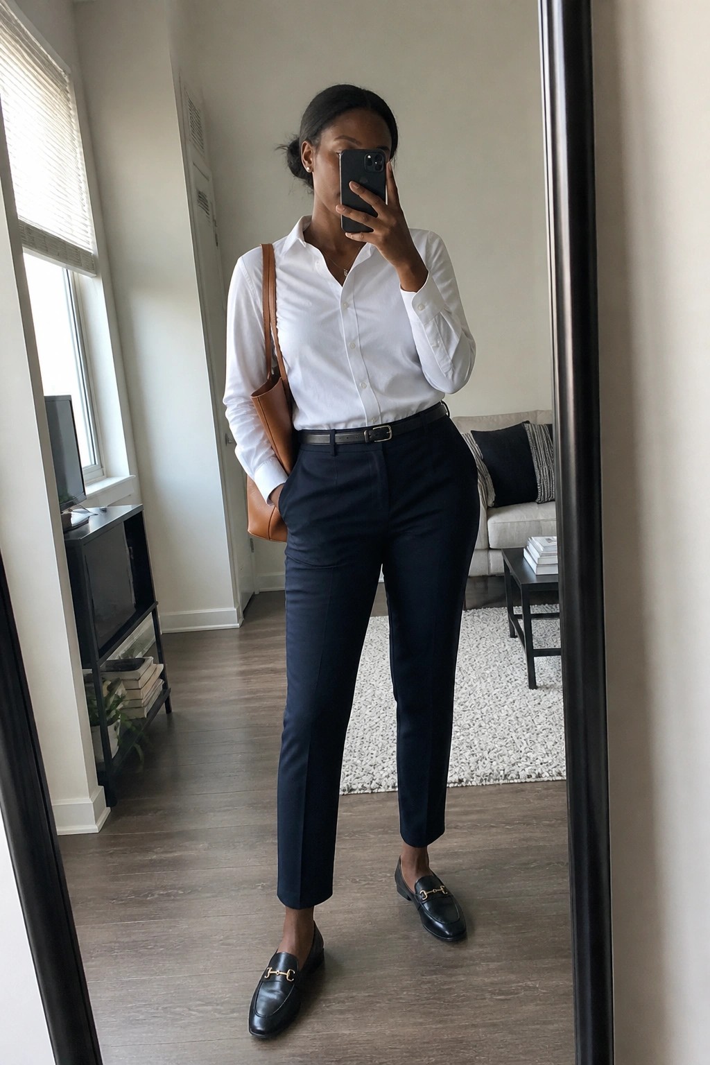 Woman in white shirt and navy pants taking mirror selfie with brown bag