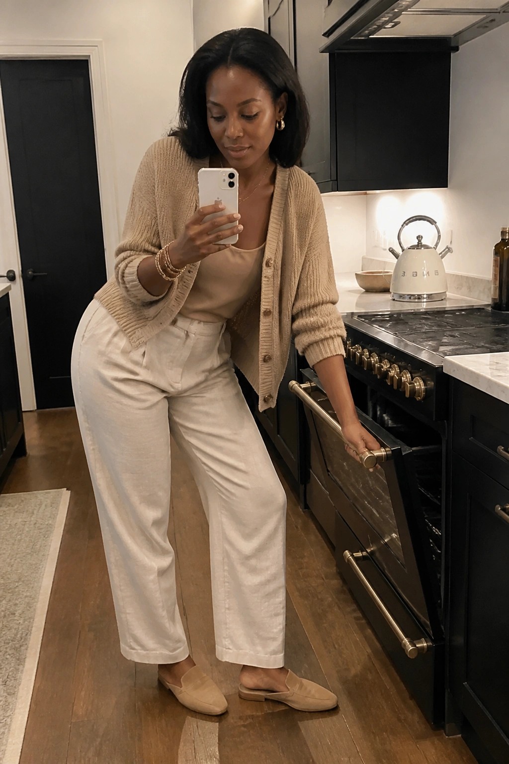 Woman in beige cardigan and white pants opening oven while taking selfie