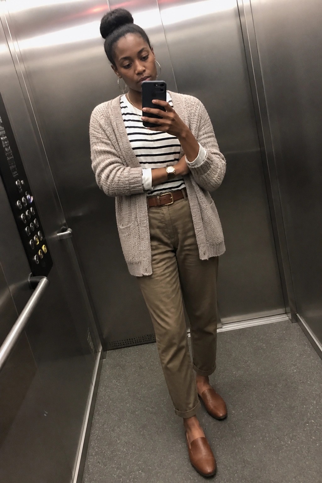 Black woman taking mirror selfie in elevator wearing open beige knit cardigan over black and white striped shirt with white cuffs showing, paired with tan chinos, brown leather belt, and matching brown loafers