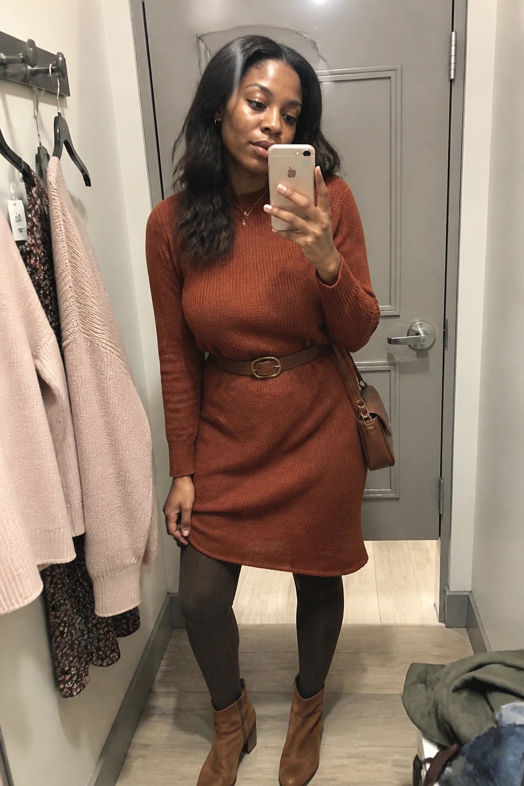 Woman wearing a rust orange knit sweater dress belted at the waist with brown leather accessories, brown tights, and suede ankle boots while taking a mirror selfie.