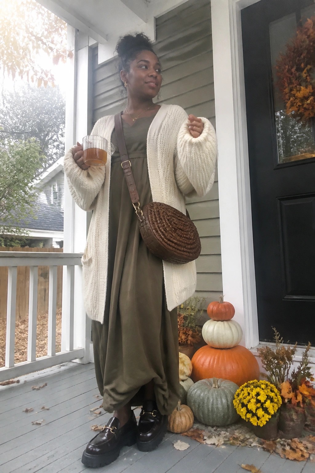 Black woman wearing oversized cream knit cardigan layered over olive green midi dress paired with brown woven crossbody bag and dark brown loafers with gold hardware.