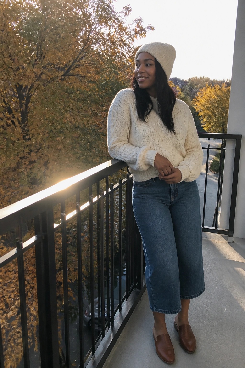 Woman in cream knit sweater and beanie on balcony with autumn trees