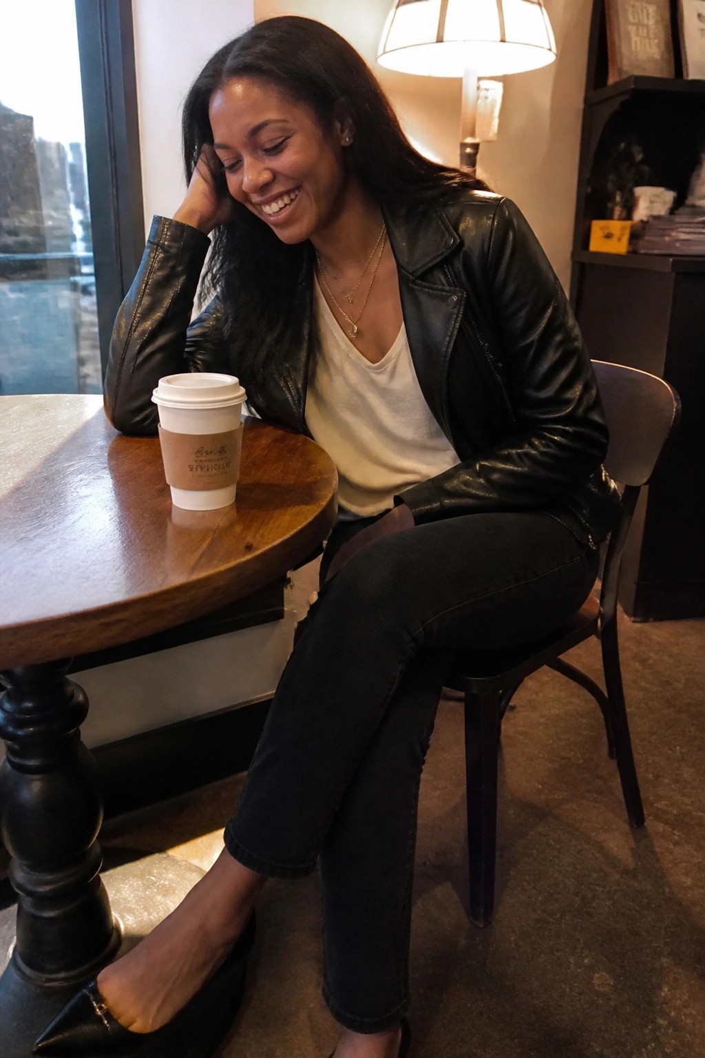 Smiling woman in black leather jacket rests hand on cheek near coffee cup