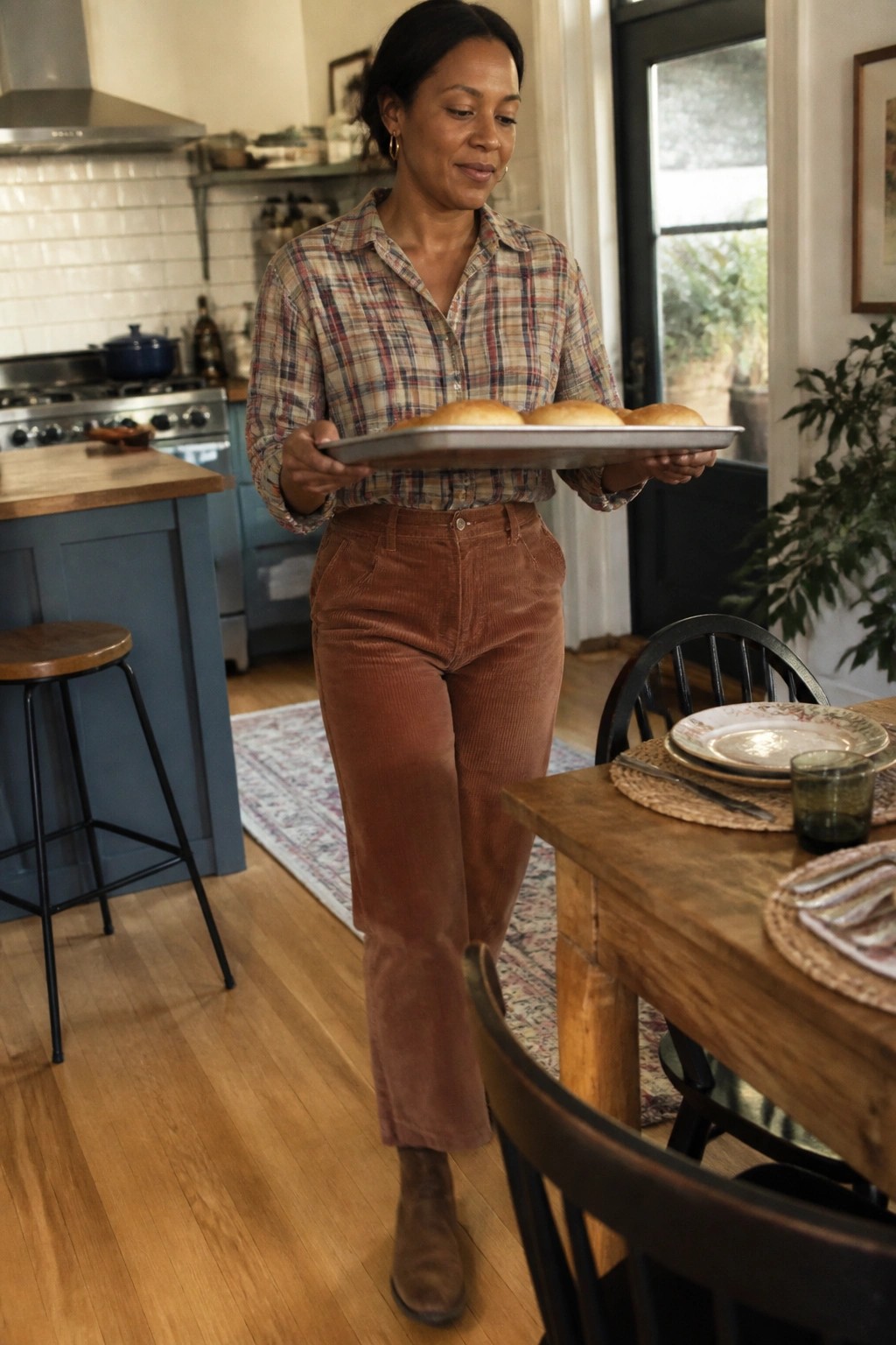 Woman wearing a multicolored plaid button down shirt tucked into rust colored corduroy pants with brown boots while holding a baking tray in a kitchen setting.