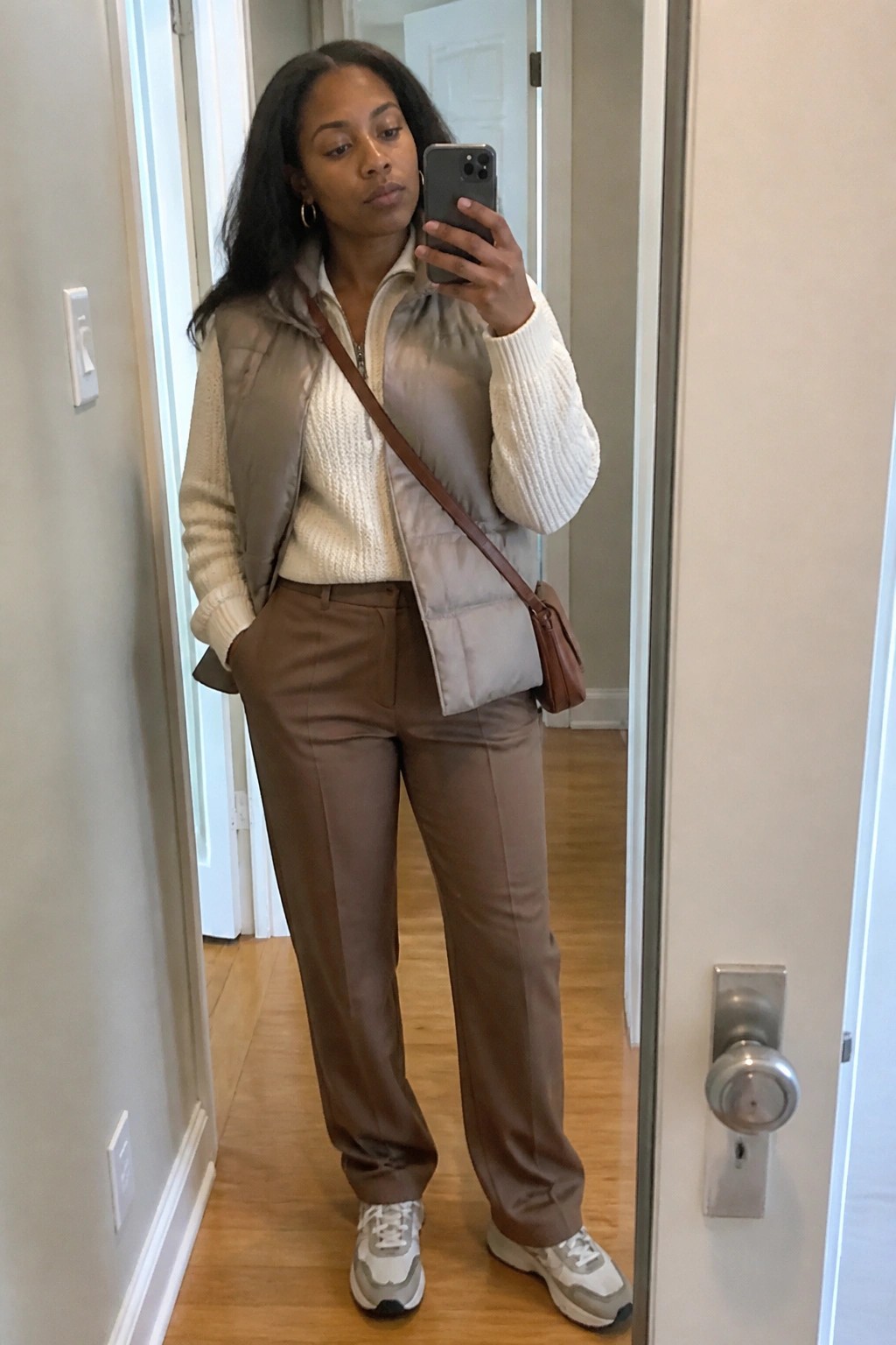 Woman in cream sweater and gray puffer vest with brown pants taking mirror selfie