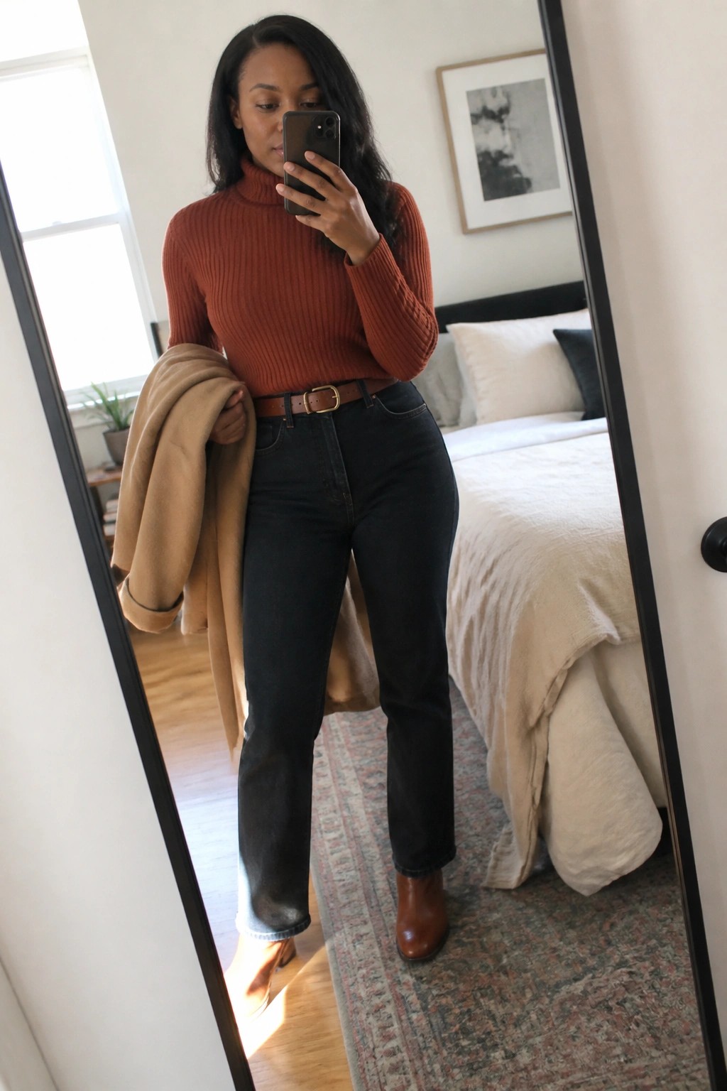 Woman in rust turtleneck, dark jeans, and brown boots holding tan coat in mirror