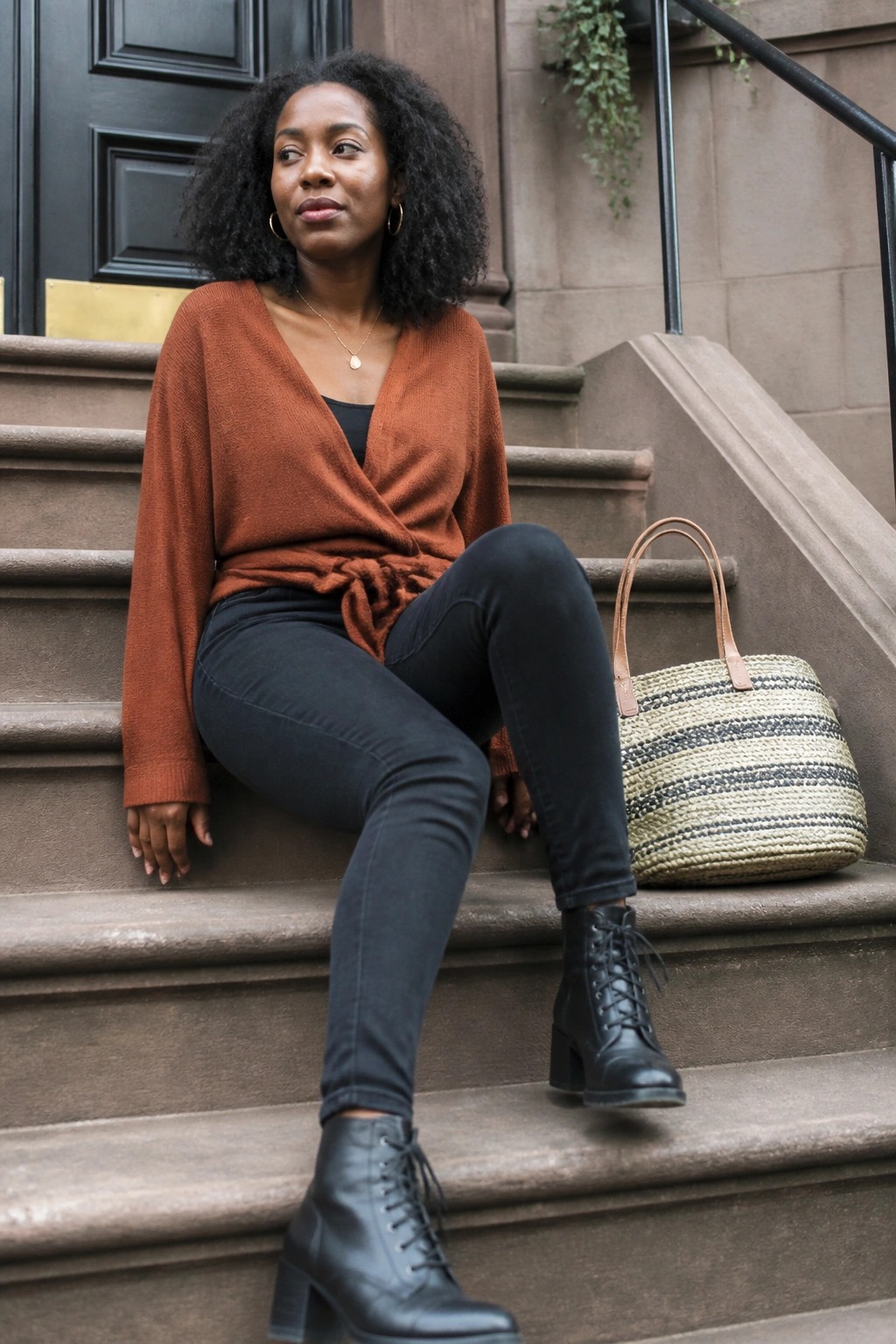 Woman in rust wrap sweater and black jeans sitting on stone steps with woven tote