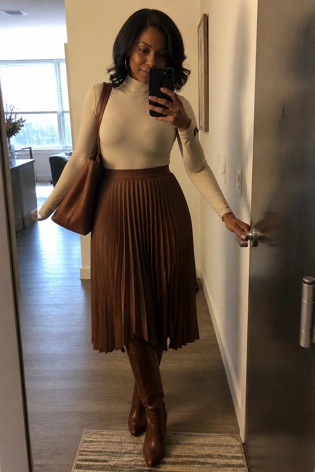 Woman in cream turtleneck and brown pleated skirt taking mirror selfie in hallway