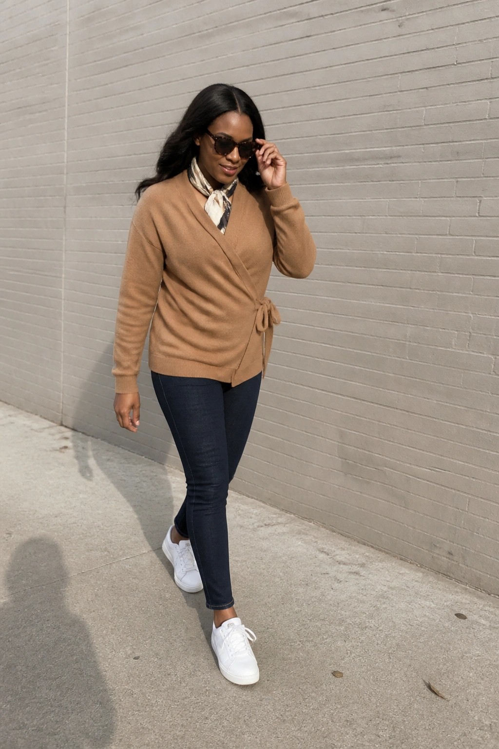 Woman in brown wrap sweater, scarf, dark jeans, and white sneakers by brick wall