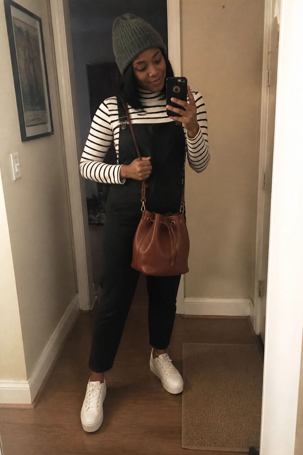 A Black woman wearing black overalls over a black and white striped turtleneck sweater paired with white sneakers a green knit beanie and a brown leather bucket bag while taking a mirror selfie.
