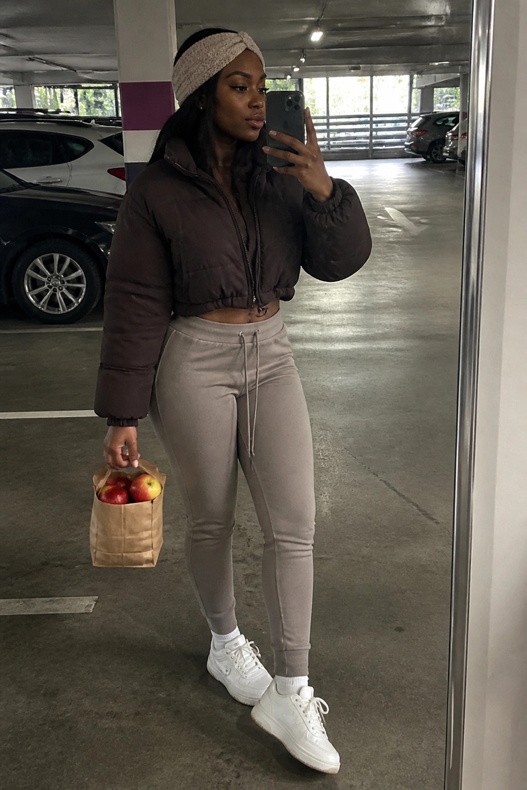 Woman in brown puffer jacket and beige joggers holding apples and phone in garage