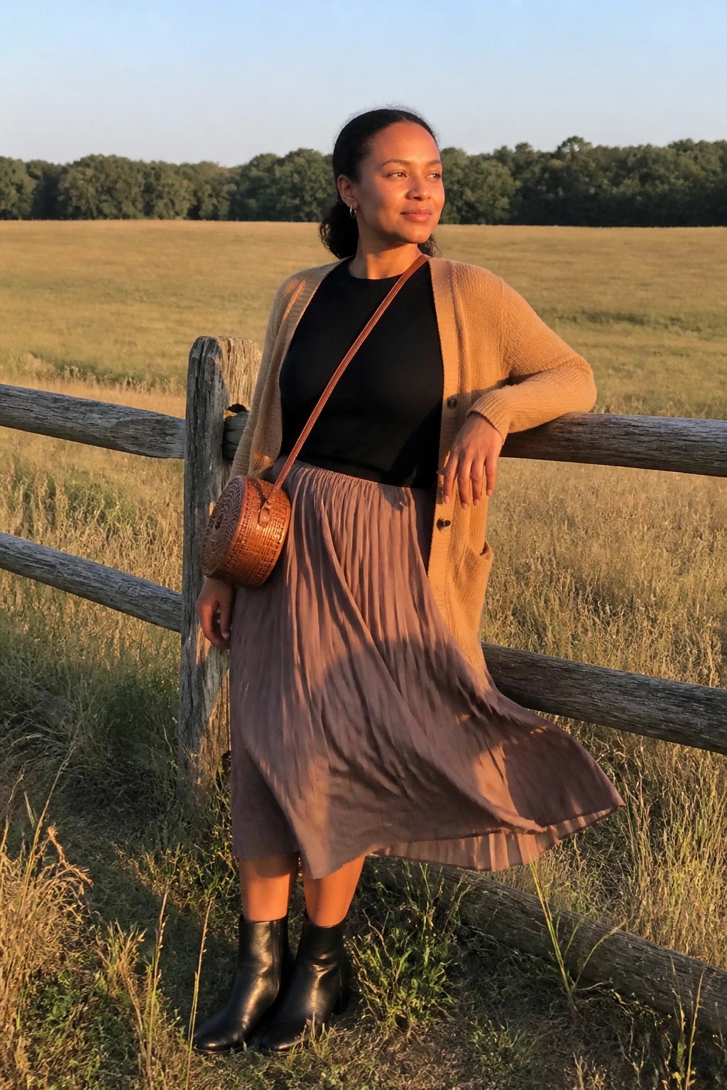 Woman in black top tan open cardigan mauve pleated midi skirt black ankle boots and round woven crossbody bag standing by wooden fence in field.