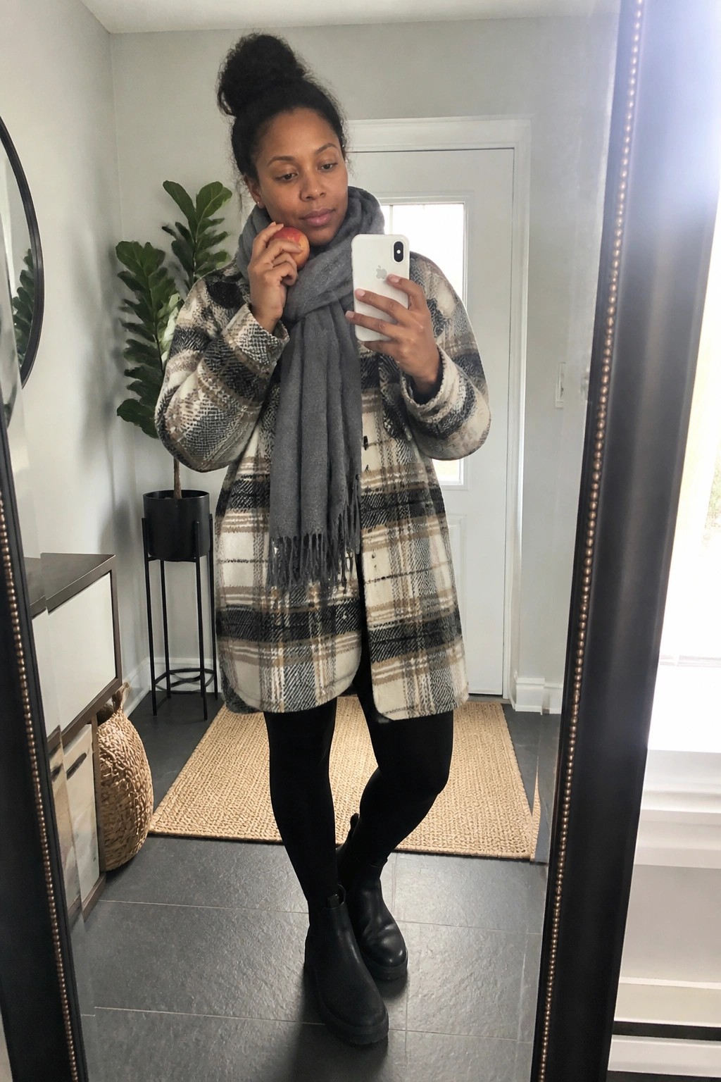 Woman in plaid coat and gray scarf taking mirror selfie while holding apple