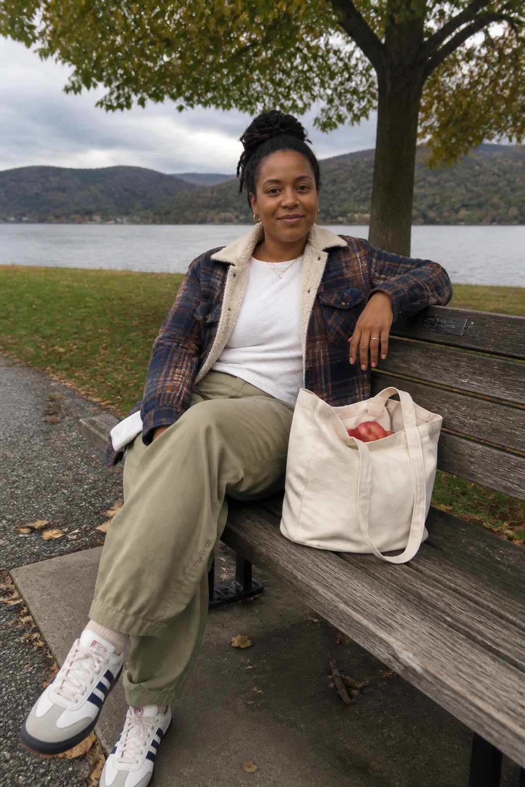 Woman in plaid sherpa jacket sits on lakeside bench beside canvas tote bag