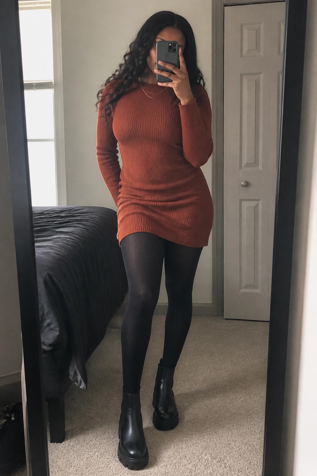 Woman in rust ribbed dress and black boots taking bedroom mirror selfie