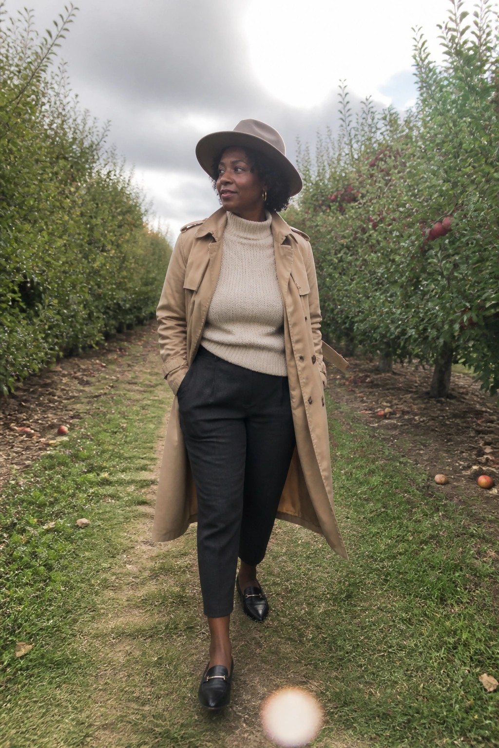 Woman in beige trench coat and hat amid apple orchard trees.