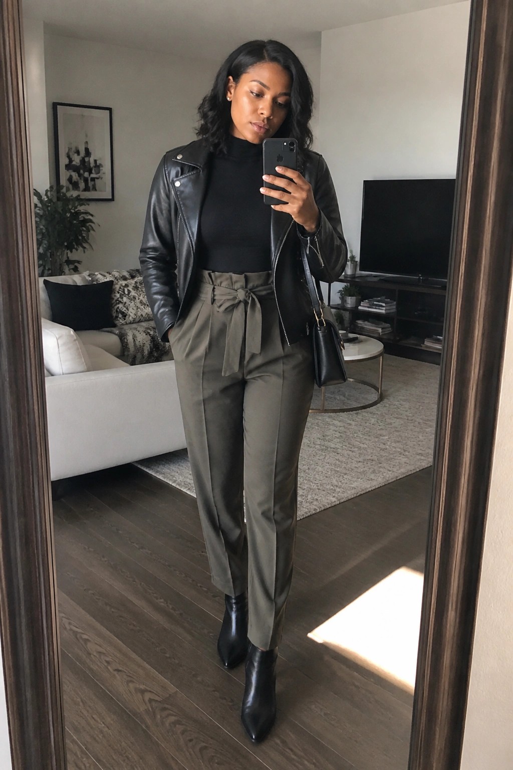 Woman in black leather jacket and olive belted pants taking mirror selfie