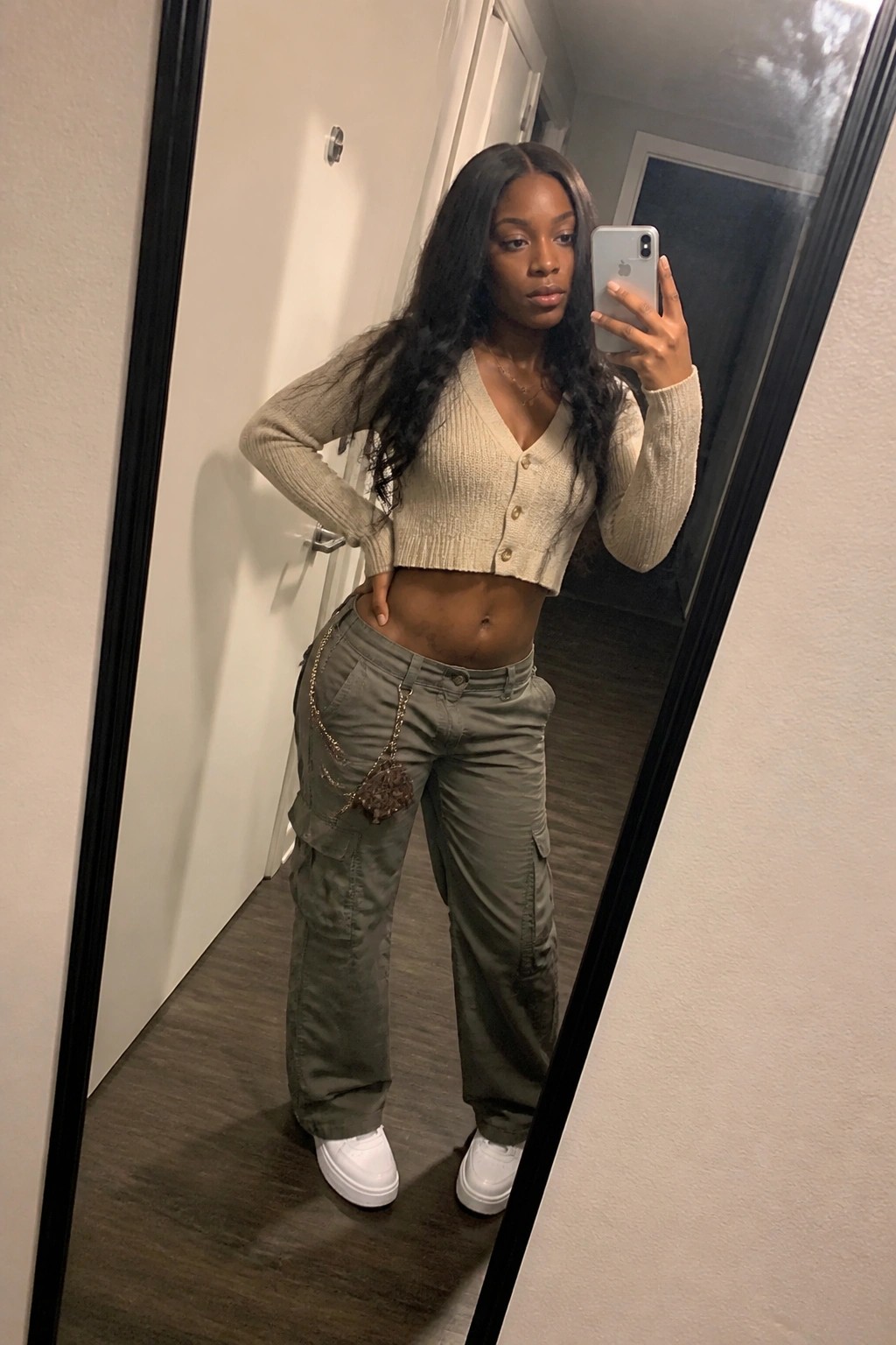Woman taking mirror selfie in cropped beige knit cardigan buttoned at the front olive green cargo pants with dangling chain accessory and white platform sneakers.