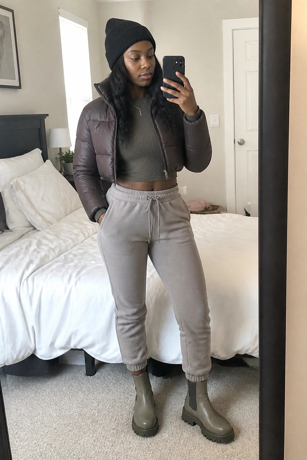 Woman in mirror selfie wearing brown cropped puffer jacket over olive ribbed crop top with taupe sweatpants, black beanie, layered gold necklace, and olive chunky ankle boots.