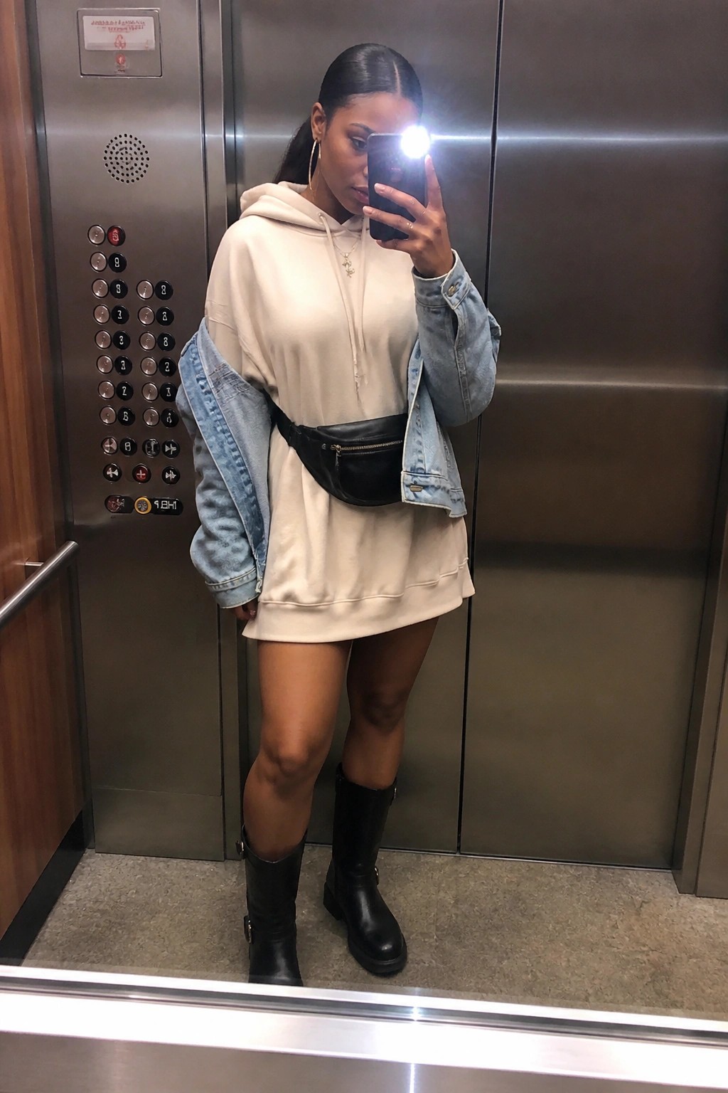 Black woman in elevator mirror selfie wearing beige hoodie dress with light denim jacket draped over one shoulder, black belt bag, hoop earrings, and black knee high boots.