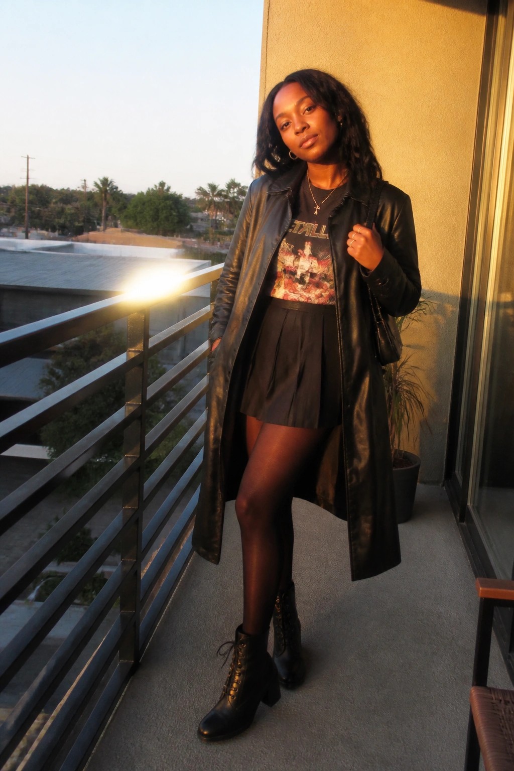 Woman in black leather coat and pleated skirt posing on sunny balcony