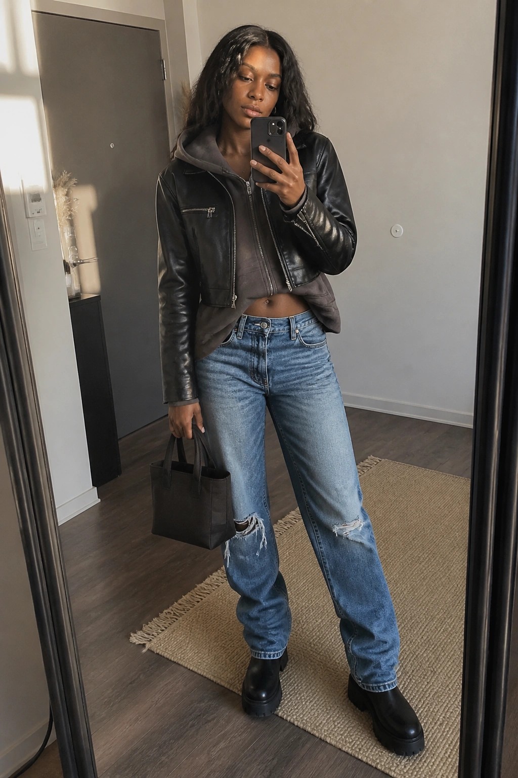 Woman in black cropped leather jacket over gray hoodie, ripped blue jeans, and black platform boots holding a small black bag during a mirror selfie.