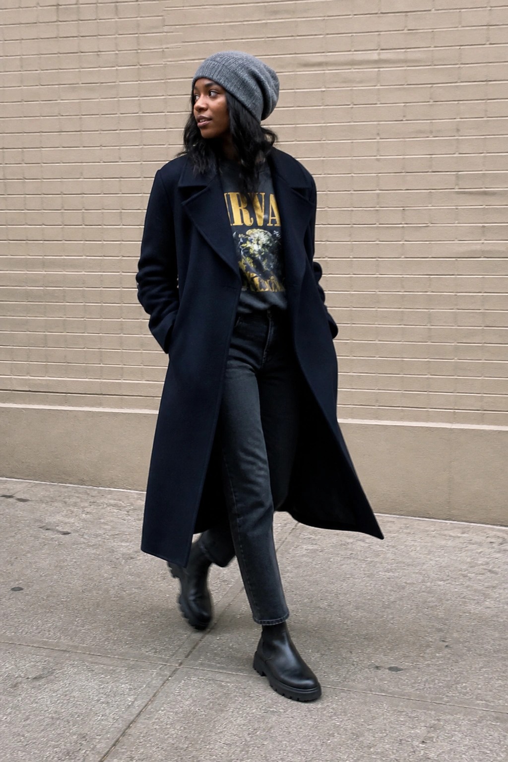 Woman in black coat over Nirvana t-shirt, grey beanie, and boots by brick wall