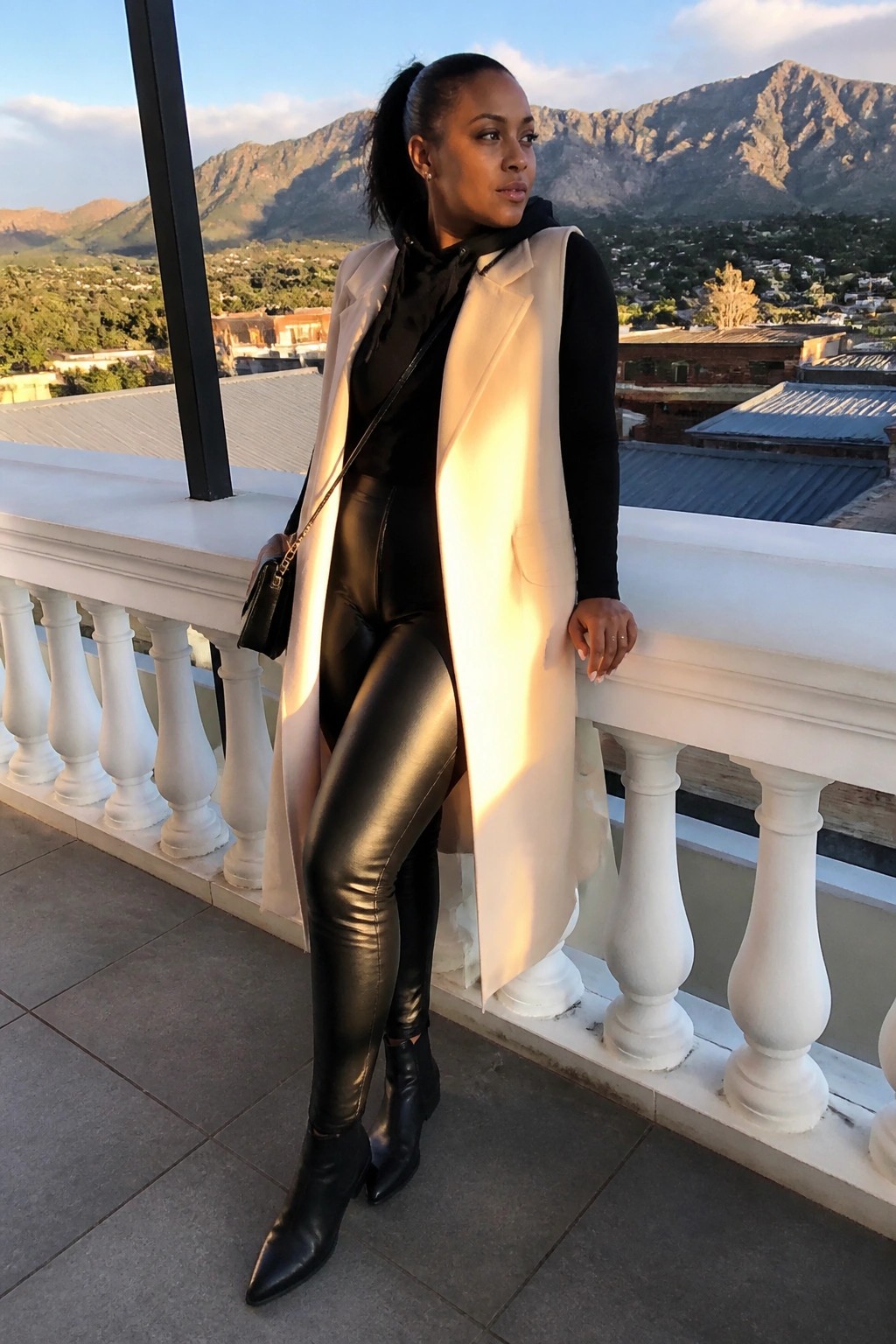 Woman wearing black bodysuit, shiny black pants, long cream sleeveless vest, black ankle boots, and small crossbody bag standing on a balcony.