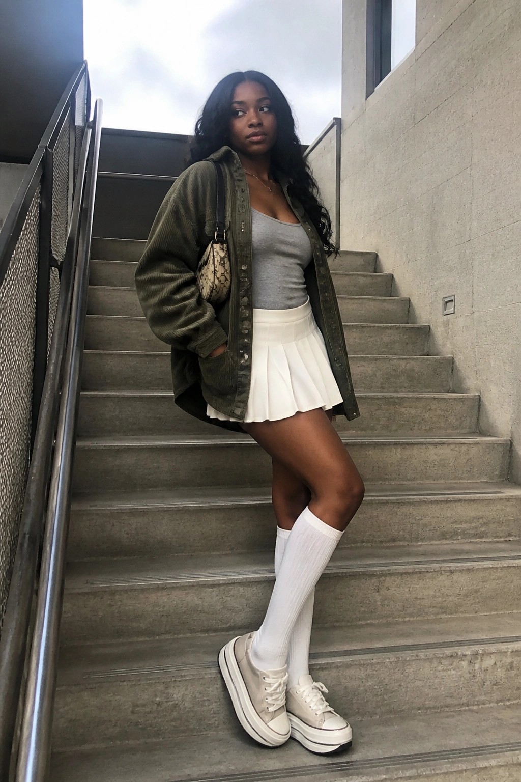Woman in green corduroy jacket, white pleated skirt, knee socks, and platform sneakers on stairs
