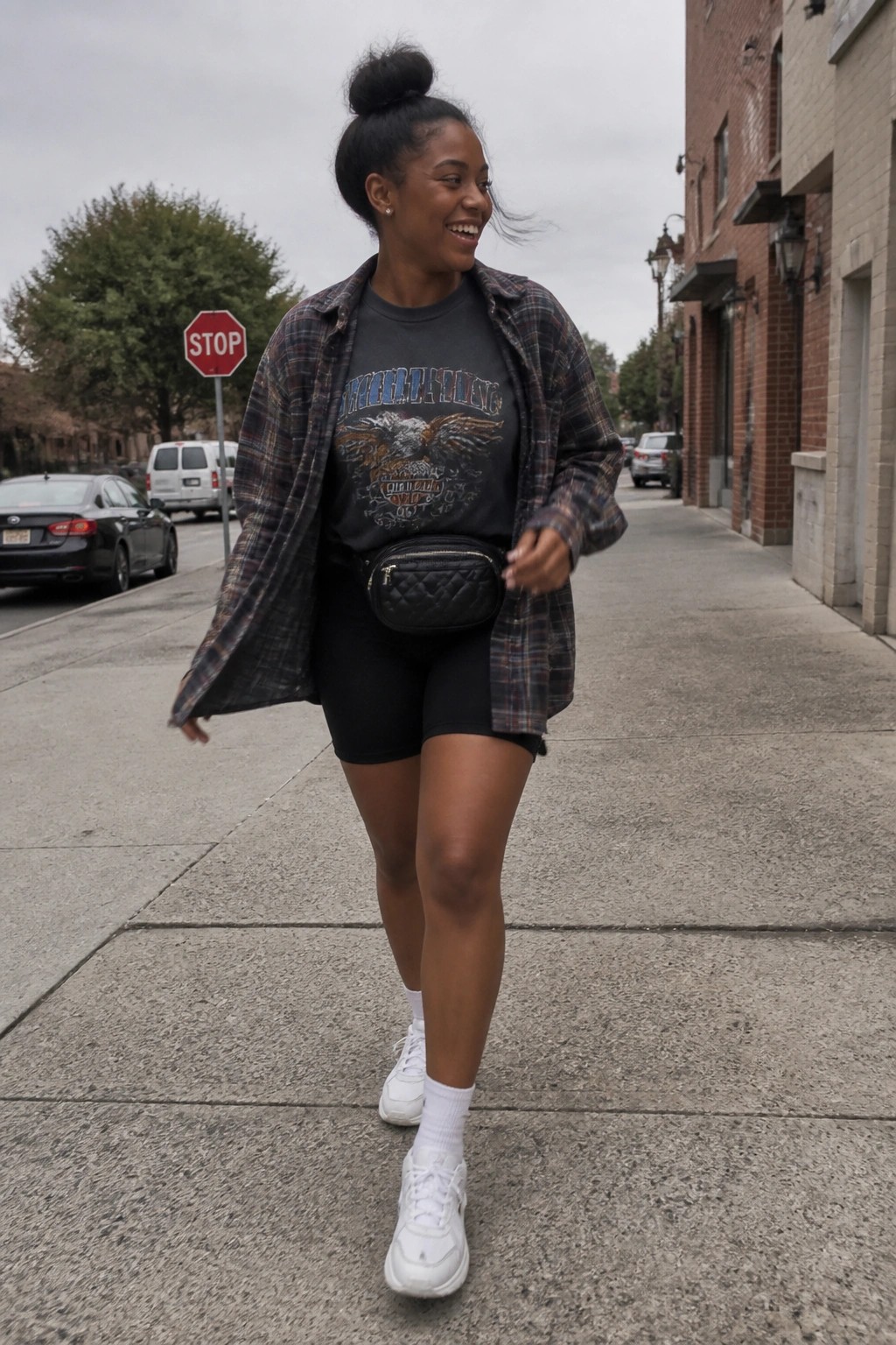 Young woman in plaid shirt and biker shorts walks smiling on urban sidewalk