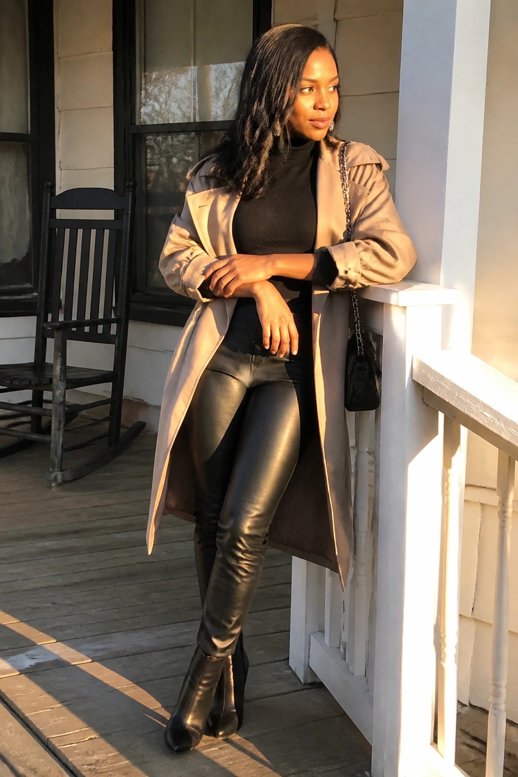 Woman in beige trench coat and black leather pants leaning on white porch railing