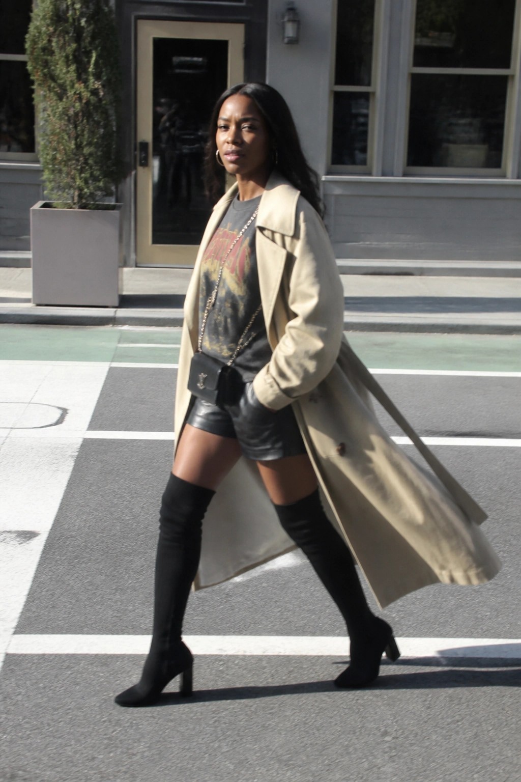 Woman in beige trench coat, leather shorts, and black thigh-high boots on city street