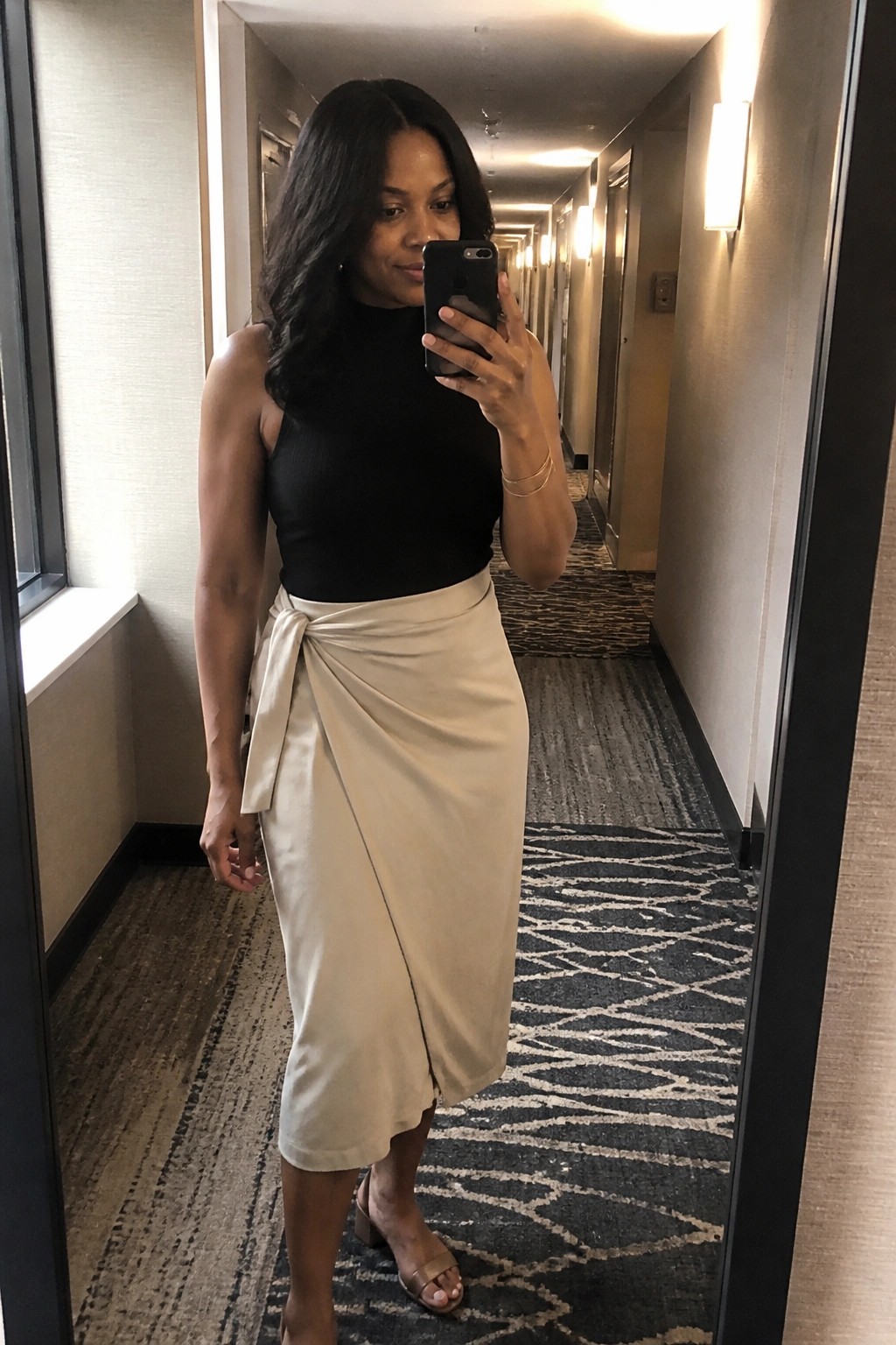 Woman in black top and beige wrap skirt taking mirror selfie in hallway