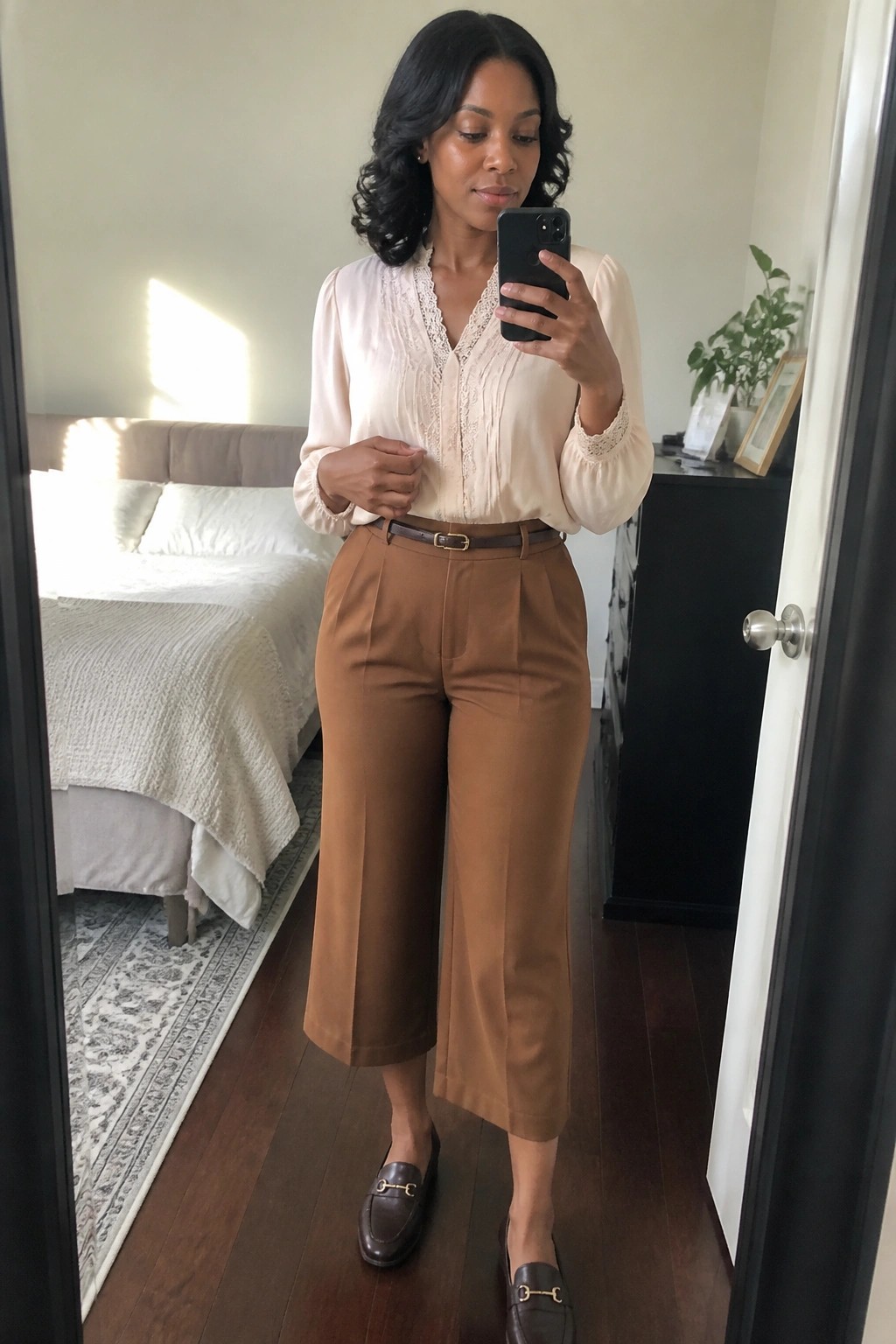 Woman in blush lace blouse and brown cropped pants taking mirror selfie