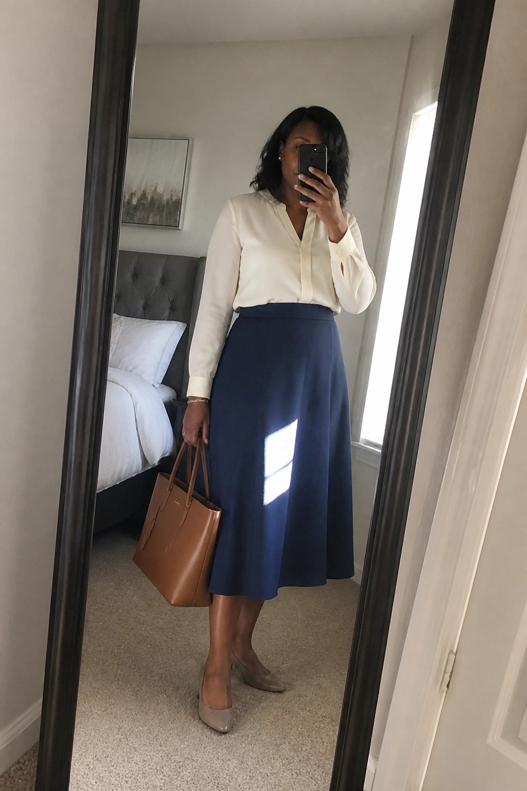 Woman in cream blouse and navy skirt holding brown bag for mirror selfie