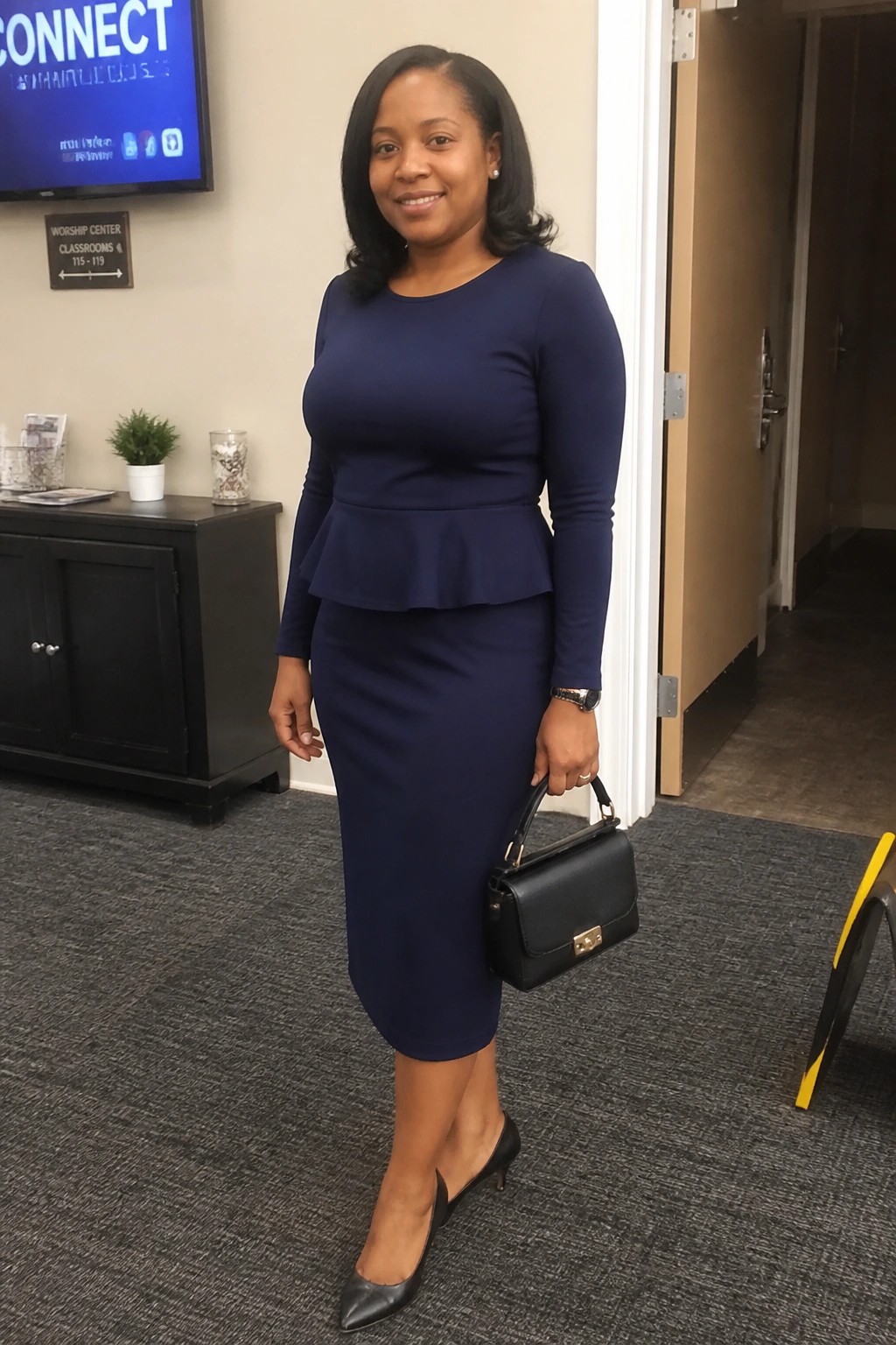 Woman in navy blue peplum top and skirt holding black handbag indoors