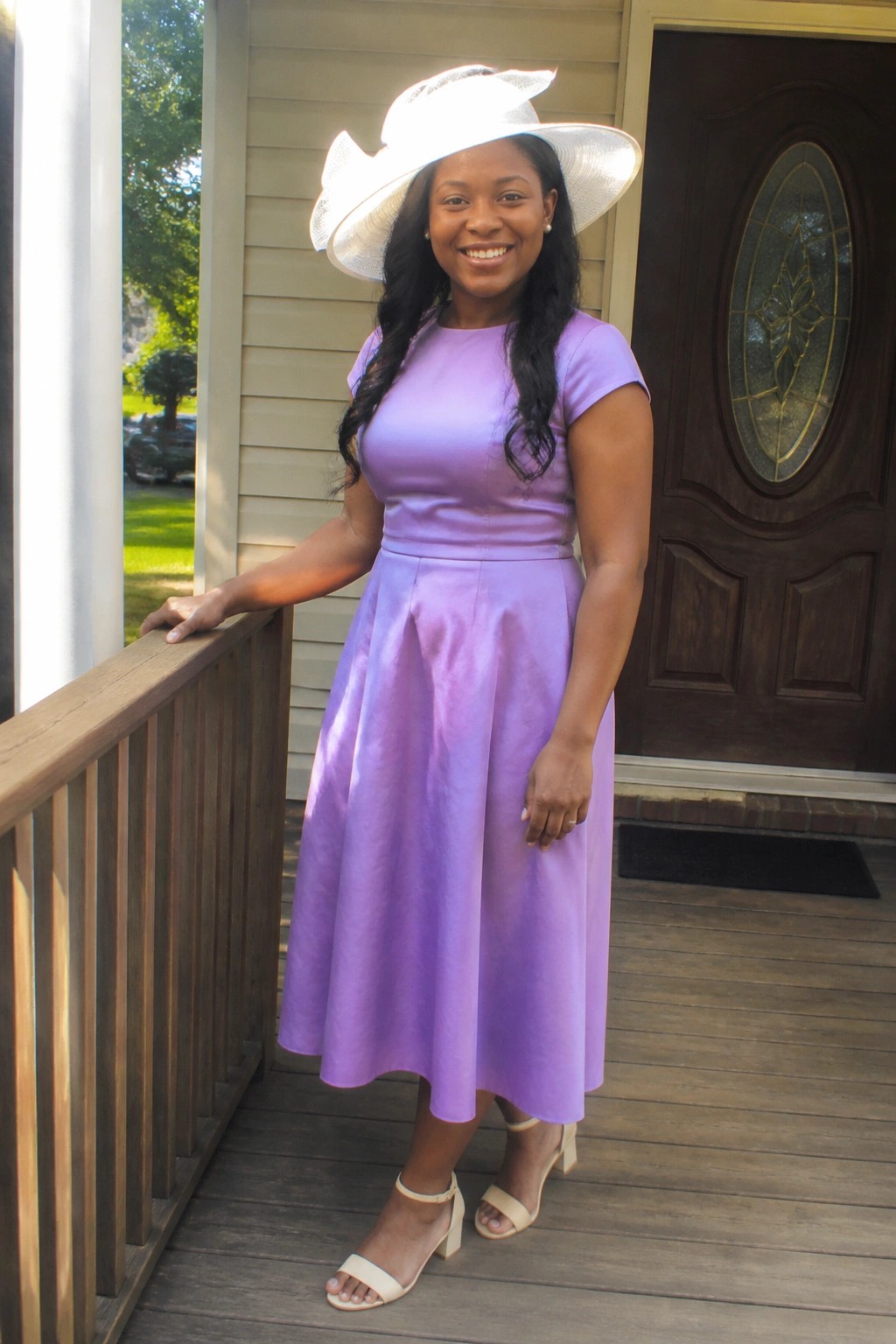 Woman wearing a lavender short sleeve midi dress with A line skirt, white wide brim hat, and beige block heel sandals standing on a wooden porch.