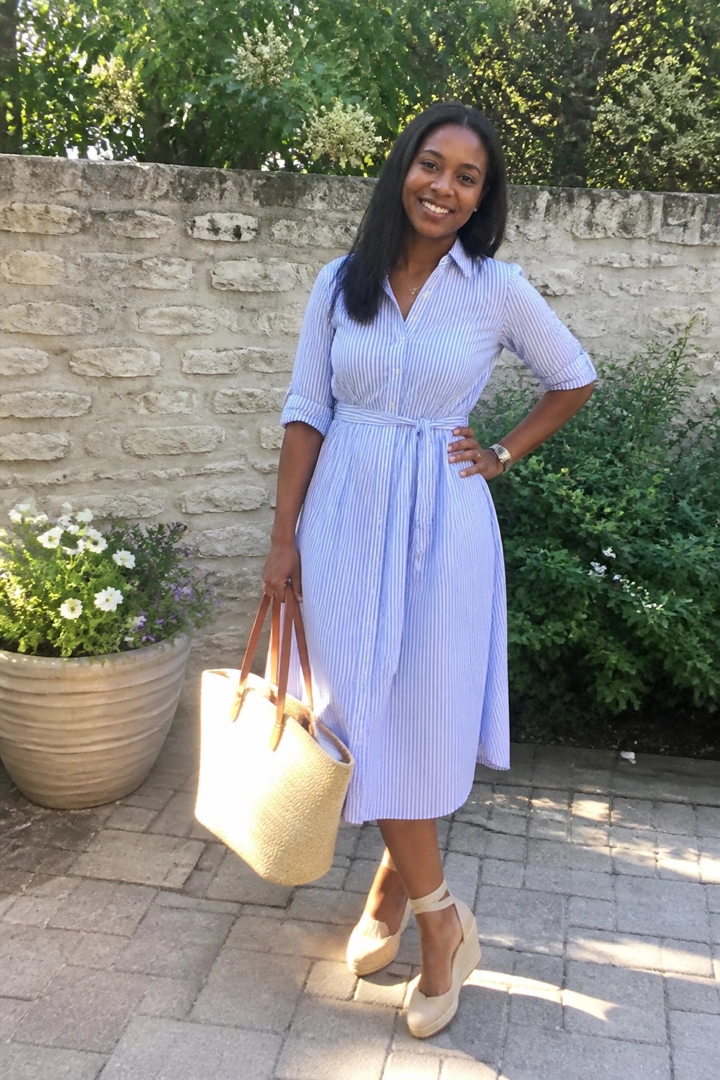 Woman in blue striped shirt dress holding straw tote by stone wall